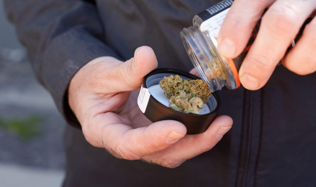 A close-up shot of a person's hands opening a premium glass jar of West Coast Cure Top Shelf Indoor Flower, preparing to extract a dense, trichome-rich cannabis bud.